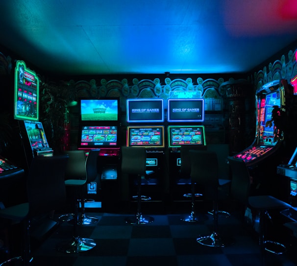 Dimly lit gaming arcade featuring a variety of brightly illuminated slot machines and video gaming screens. Neon signs and colorful displays create a vibrant atmosphere. The arrangement of gaming machines suggests an inviting space for entertainment, with chairs positioned for players.