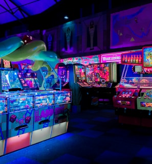 An indoor arcade filled with vibrant gaming machines lit with neon blue and pink lights. The scene includes various arcade games with flashing screens and colorful displays, and a large overhead structure resembling a green snake. The background shows illuminated signs and themed decor, contributing to an immersive entertainment atmosphere.