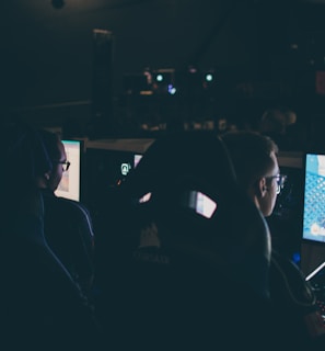 Several individuals are sitting in front of computer screens in a dimly lit room, likely engaging in gaming or working on tasks. The glow of the monitors illuminates their faces, creating a focused and immersive atmosphere. The surroundings suggest a setting like a gaming event or a LAN party.