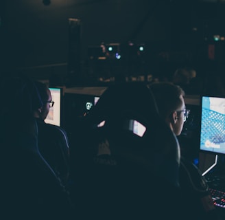 Several individuals are sitting in front of computer screens in a dimly lit room, likely engaging in gaming or working on tasks. The glow of the monitors illuminates their faces, creating a focused and immersive atmosphere. The surroundings suggest a setting like a gaming event or a LAN party.