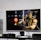 A flat-screen TV displays a video game interface featuring a character from Mortal Kombat in the center. Beside the TV is a stack of video game cases for the PlayStation 3, including titles like FIFA 14 and Grand Theft Auto. Below the TV, there is a gaming console, controllers, and other gaming equipment.