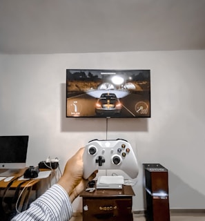 A person is holding a gaming controller in front of a flat-screen television displaying a racing video game. The room has a computer setup on a wooden desk with cables, a desktop computer monitor, and other accessories. The focus is on the interaction between the person and the gaming environment.