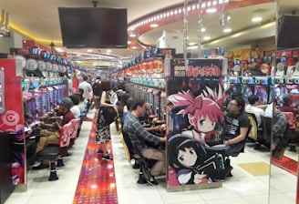A bustling arcade filled with people playing various gaming machines. Brightly colored displays and animated characters are prominent on posters and screens. The atmosphere is lively with patrons seated in rows, focusing on the games. Overhead signage and electronic scoreboards are visible, contributing to the busy and vibrant environment.