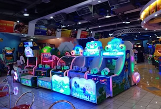 A brightly lit arcade filled with colorful gaming machines sits under ceiling lights. Neon lights illuminate various game stations, creating a playful and vibrant environment. A few red chairs are scattered around the reflective, tiled floor, and a person is observing one of the machines. The atmosphere is lively and cheerful, featuring games with cartoonish designs and engaging themes.