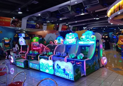 A brightly lit arcade filled with colorful gaming machines sits under ceiling lights. Neon lights illuminate various game stations, creating a playful and vibrant environment. A few red chairs are scattered around the reflective, tiled floor, and a person is observing one of the machines. The atmosphere is lively and cheerful, featuring games with cartoonish designs and engaging themes.