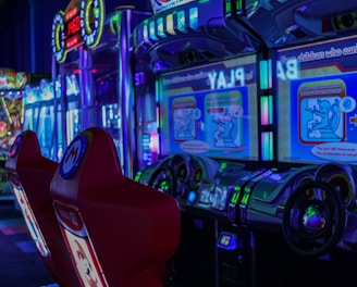 A vibrant arcade environment with several illuminated gaming machines. The image features arcade racing games with visible steering wheels, colorful control panels, and screens displaying game instructions. Bright LED lights in various colors illuminate the machines and surrounding area.