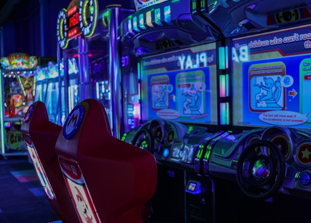A vibrant arcade environment with several illuminated gaming machines. The image features arcade racing games with visible steering wheels, colorful control panels, and screens displaying game instructions. Bright LED lights in various colors illuminate the machines and surrounding area.